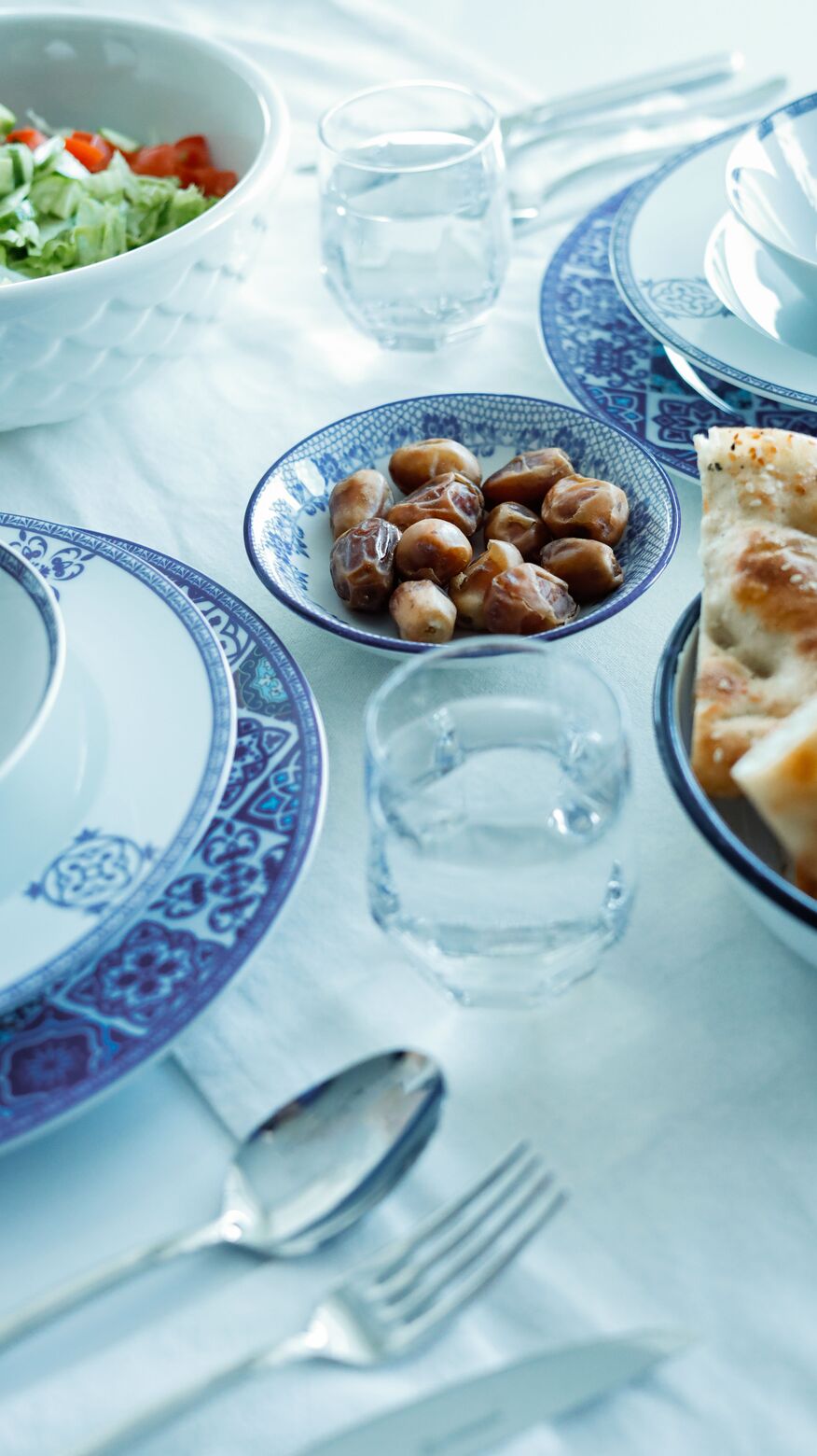Blue and white patterned plates with dates, glasses of water and cutlery on a light iftar table