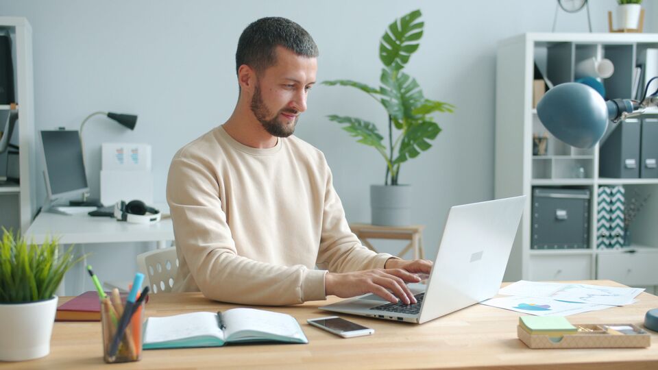 Smiling man working on a laptop at a bright home desk with plants and shelves